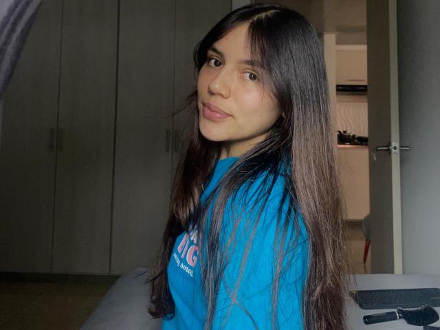 Photograph of SophiaMarx with long, straight black hair, wearing a blue shirt, looking over her shoulder with a slight smile. Background includes a dark wardrobe and a partially open door. She has a sexy, confident vibe.