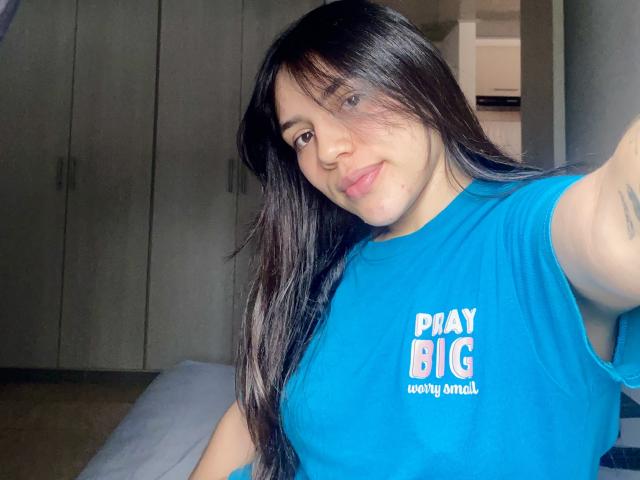 Fotografia de SophiaMarx, uma mulher curvilínea com cabelos longos e pretos, vestindo uma camiseta azul vibrante com a inscrição "PAY BIG WONG DONG". texto. Ela está tirando uma selfie, com o braço estendido, exibindo seus seios grandes..