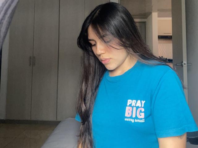 Fotografie von Sophia Marx, einer jungen Frau mit langen, schwarzen Haaren, die ein hellblaues T-Shirt mit dem Aufdruck "PAY BIG" trägt. Sie blickt nach unten und zeigt dabei ihr weiches, rundes Gesicht und ihr dezentes Make-up..