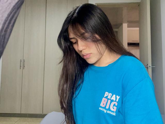 Foto van SophiaMarx met lang, recht zwart haar, die een felblauwe blouse met witte tekst draagt en naar beneden kijkt. Een licht verleidelijke uitdrukking. Achtergrond van de slaapkamer, lichte kasten. Seksuele spanning..