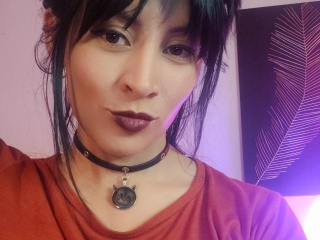 Close-up of IssisDiosa, Asian woman with pale skin, black hair in a messy bun, wearing a red shirt and black choker with a cat pendant. Sexual tension, slightly smug expression.