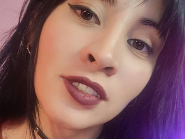 Close-up selfie of IssisDiosa, an Asian woman with fair skin, dark brown eyes, and black lipstick. She has dark hair and is looking directly at the camera with a slight smirk. Her expression is sexy and seductive.