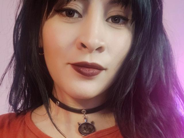 Close-up of IssisDiosa with long black hair, fair skin, dark lipstick, and a black choker with a pendant. She gazes directly at the camera with a subtle, confident smirk.
