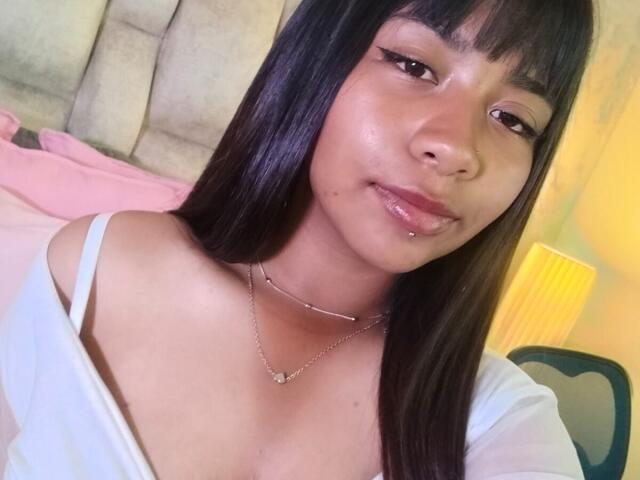 Close-up selfie of NahomyNoir, a young woman with straight black hair, bangs, and a slightly shiny face, wearing a white off-shoulder top, looking seductively at the camera, lips slightly parted.