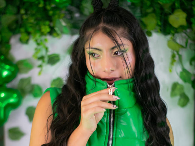 LeiaLaurent, met lang zwart haar, draagt een groene latex top met rits, groene oogschaduw en een ring, en ze bijt in de rits. terwijl je recht in de camera kijkt. Groene bladeren op de achtergrond.