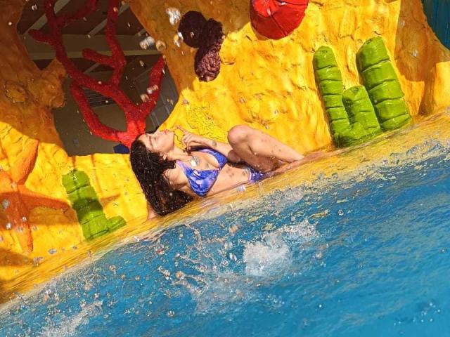 Digital artwork of LoreneCurly with wet black hair, blue bikini, leaning against a yellow rock wall with green boulders. Water splashes around, creating a sexual tension vibe. Bright colors, vivid textures.