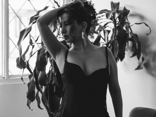 Photographie en noir et blanc de Samantha Antolin, debout, une main dans ses cheveux bouclés, portant une robe à décolleté plongeant et sans bretelles. robe noire, ombragée par une grande plante à feuilles, intense, sexy, éclairage sombre et suggestif..