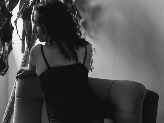 Schwarz-weißes Foto von Samantha Antolin, Rückenansicht, sie lehnt sich an ein Sofa, lockiges Haar, schwarzes Top ohne Träger, sexuell ansprechend. Spannung, schattiges Gesicht, Pflanzenblätter im Hintergrund, stimmungsvolle Beleuchtung..