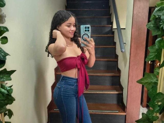 LoreneCurly se toma un selfie en un rellano de escaleras, vistiendo una blusa roja sin tirantes, atada al frente, y jeans azules de cintura alta. Ella tiene el pelo negro y rizado, y se señala a sí misma. Increíblemente sexy..