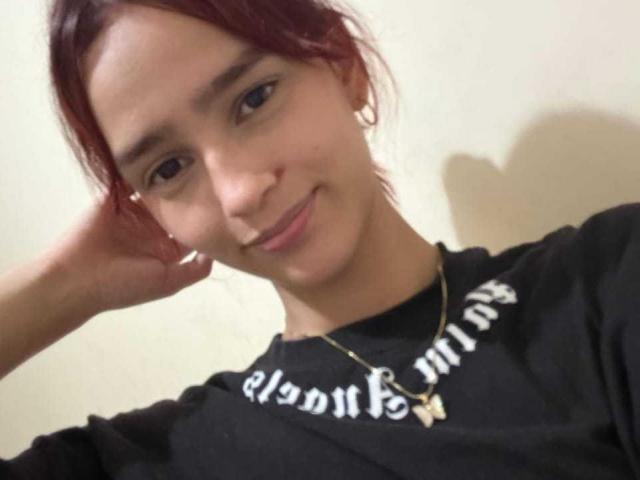 Fotografía de Melody Mansson con cabello castaño rojizo, sonriendo ligeramente, con la mano apoyada en la cabeza, vistiendo una sudadera negra. Con texto en blanco y un collar dorado con un dije de cruz. Qué lindo/a..