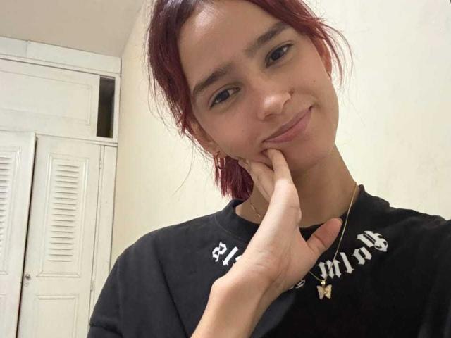 Fotografia di Melody Mansson con i capelli rossi corti, che sorride leggermente e che tocca il mento con una mano. Indossa una maglietta nera con scritte bianche. Sullo sfondo, si vedono porte bianche con persiane. Casual, leggermente sensuale.