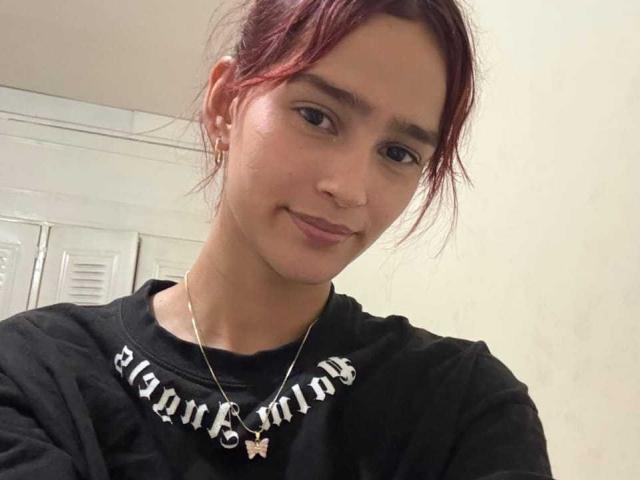 Fotografía de Melody Mansson con cabello castaño rojizo, vistiendo una camisa negra con la palabra "Bergman" y un collar de plata, sonriendo. ligeramente.
el fondo tiene taquillas blancas. MelodyMansson tiene una vibra sutil, sexy y relajada..