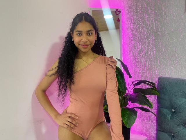 EmaFfox, met lang, krullend zwart haar, staat zelfverzekerd in een roze, eenschoudrig lycra pak, met haar hand op haar heup, en er schijnt een roze neonlicht. achtergrond, groene plant aan de rechterkant, grijze bank gedeeltelijk zichtbaar.