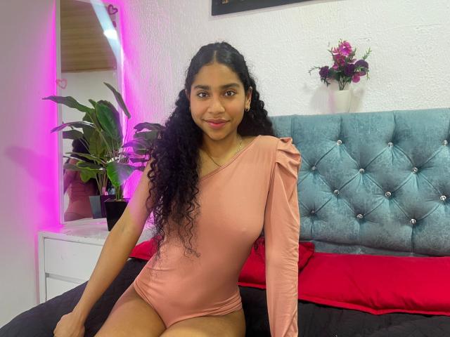 EmaFfox está sentada em uma cama, vestindo um macacão rosa com uma alça, e tem longos cabelos cacheados pretos. Suas pernas estão abertas, exibindo suas coxas tonificadas. Uma cabeceira azul com textura e almofadas vermelhas estão atrás dela..