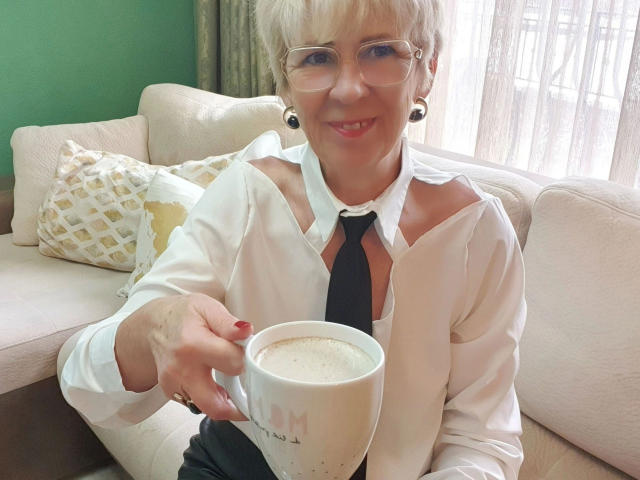 OOHotEveOo-sex con cabello corto y gris, gafas y aretes negros, vistiendo una camisa blanca desabrochada y una corbata negra, sosteniendo una taza blanca, sentado en un sofá color crema, sonriendo. Luz brillante que entra por la ventana.