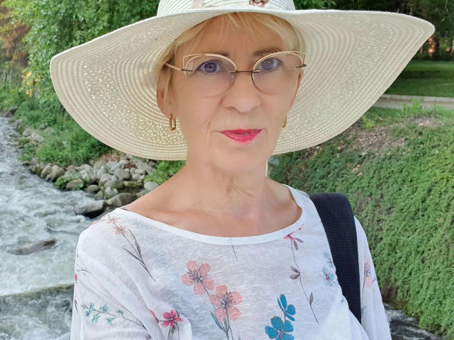 OOHotEveOo-sex, una mujer mayor con cabello rubio corto, que lleva un sombrero blanco de ala ancha, gafas redondas y lápiz labial rojo, camisa con estampado floral, correa negra, de pie junto a un río que fluye, fondo de vegetación. Muy atractivo..