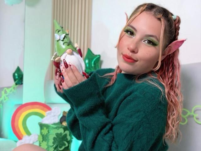 DominicTheOne, with elf ears, holds a white dildo, wearing a dark green sweater, pink hair, red lipstick, and green eyeshadow, sitting amidst a colorful, rainbow-themed background. Fucking cute.