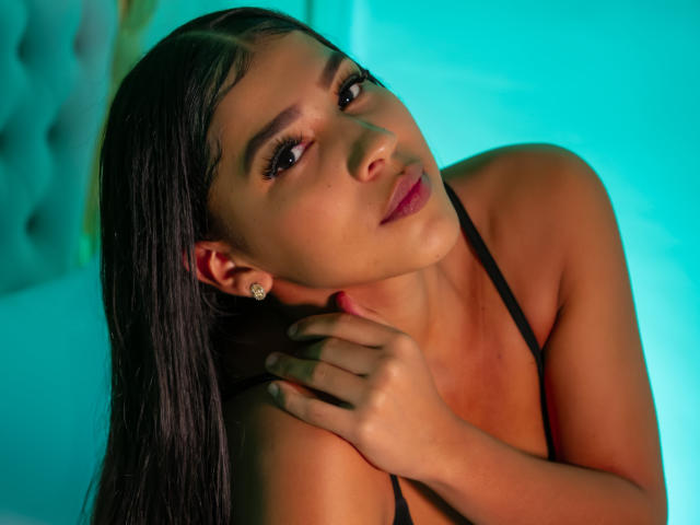 Fotografie von BiancaJanes, Kopf leicht nach hinten geneigt, dunkles, nasses Haar, intensiver Blick, Hand am Hals, schwarzes Top mit Trägern, leuchtend blau. Hintergrund, Schattenspiel im Gesicht, sexy, verführerisch, selbstbewusster Ausdruck..