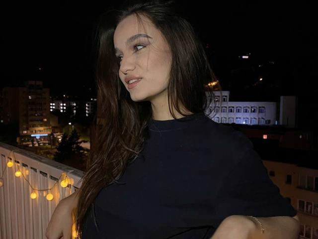 Fotografía nocturna de LenaLaurent, una mujer morena con cabello largo, vestida con una camisa negra, de pie en un balcón con una barandilla. luces, mirando hacia un lado con una ligera sonrisa. Paisaje urbano en el fondo.