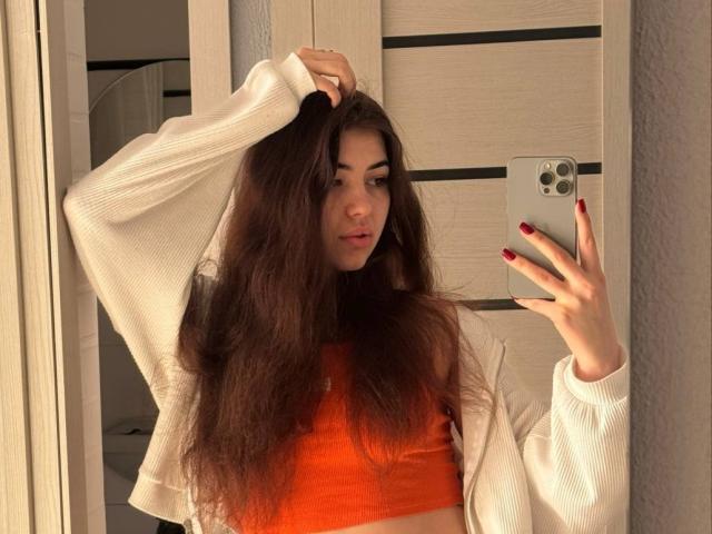 Clarinda Wiatr, com cabelos castanhos longos e desalinhados, vestindo um suéter branco por cima de uma cropped vermelha, segurando um iPhone vermelho. unhas, posando em frente a uma parede de madeira, mão no cabelo.