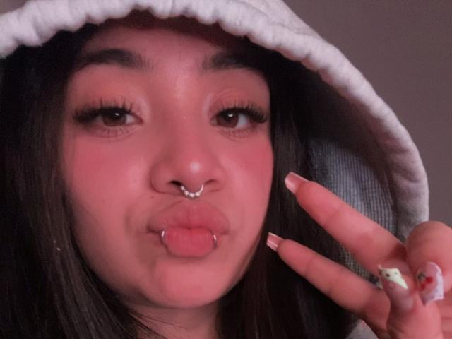 Selfie de perto de AmaraKazumi, com a pele levemente rosada, cabelo escuro e um capuz branco. Ela está fazendo um sinal de paz com a língua de fora, usando um piercing no nariz e um anel. Muito quente.