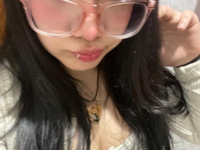 Close-up selfie of AmaraKazumi with black hair, pink-tinted glasses, and a lip piercing. She's touching her glasses with one hand, wearing a white top and a necklace. Fucking hot.