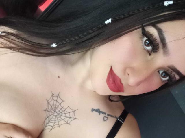 Foto ravvicinata di Chloey69, con capelli scuri raccolti in trecce, eyeliner nero intenso, rossetto rosso, top nero con spalline e un disegno di ragnatela. Tatuaggio sul petto e un piccolo tatuaggio a forma di cuore vicino alla clavicola. Un aspetto incredibilmente attraente..