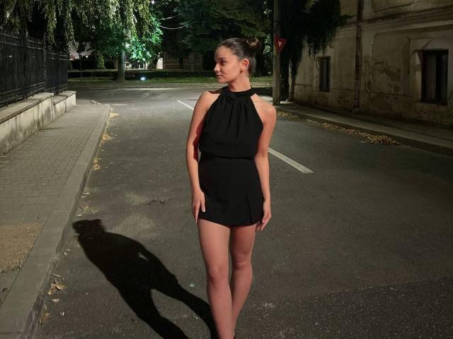 LenaLaurent está em uma rua escura e vazia à noite, vestindo um mini-vestido preto sem mangas e com alças. A sombra dela é longa, e ela desvia o olhar com uma expressão séria. Ambiente urbano.