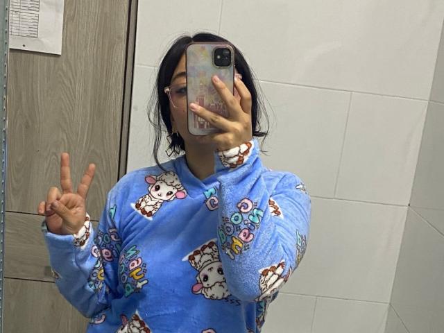 AmaraKazumi takes a selfie, face obscured by phone, wearing a blue Hello Kitty-patterned shirt. She flashes a peace sign with her right hand. Her black hair is messy.
