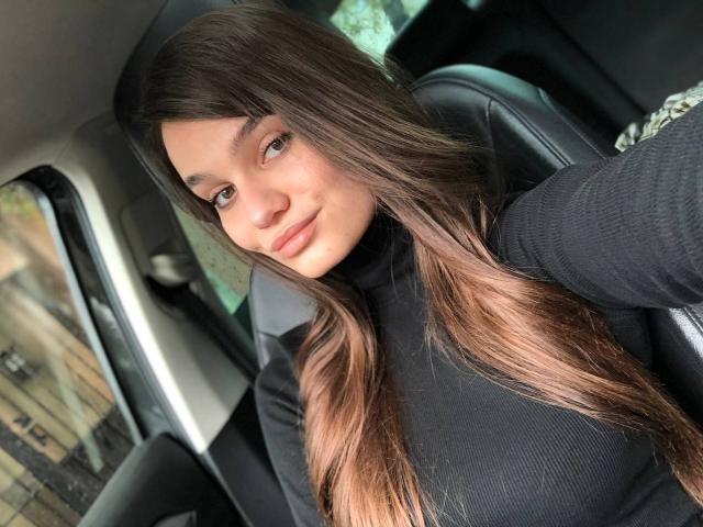 Fotografia di LenaLaurent all'interno di un'auto, che si sporge in avanti con lunghi capelli castani, indossa una maglietta nera, occhi azzurri che guardano. la fotocamera, luce soffusa, sfondo leggermente sfocato, atmosfera di un selfie spontaneo..