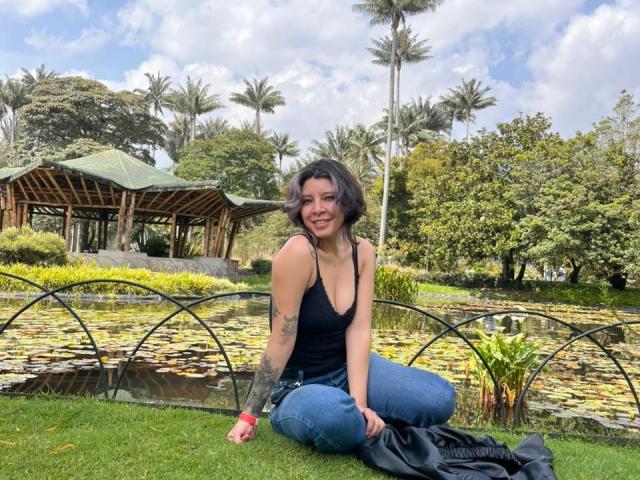 DarckVelvet, a brunette woman with a tattoo on her left arm, sits on grass by a pond, wearing a black tank top and blue jeans. Palm trees and a wooden pavilion in the background. Fucking relaxed, smiling, with a black cloth beside her.