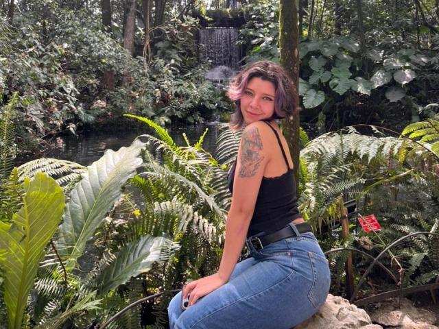 DarckVelvet kneels in lush, green foliage, wearing a tight black tank top and high-waisted jeans, showcasing a large tattoo on her upper arm. She has dark hair and a seductive look.