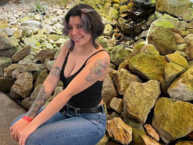 DarckVelvet, a tattooed woman with short black hair, crouches in front of a rocky wall, wearing a black tank top and blue jeans, smiling seductively, displaying visible cleavage. Bright outdoor lighting.