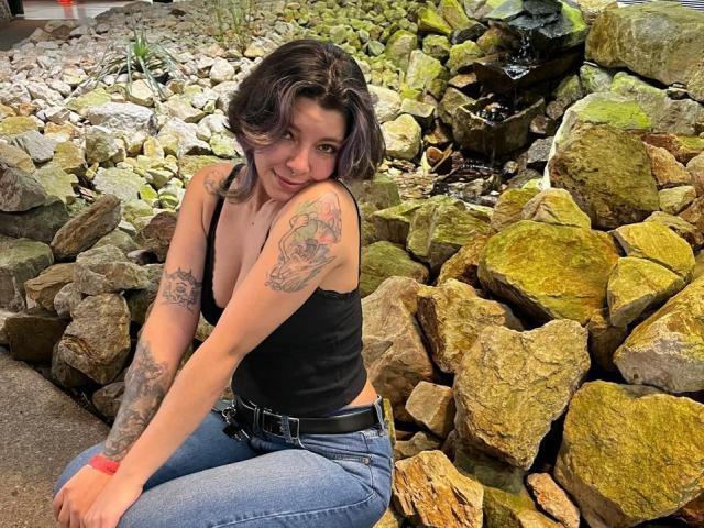 DarckVelvet knielt op de grond, omringd door grote, met mos bedekte rotsen. Ze heeft tatoeages op beide armen, draagt een zwart tanktopje en blauwe jeans, met een subtiele, sexy glimlach..