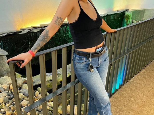 DarckVelvet, lean against black metal railing, wearing black tank top, blue jeans, belt with keys, red bracelet, tattooed arm. Sexual tension, leaning slightly forward, cock visible through jeans.