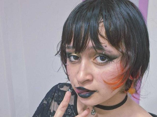 Close-up photo of MichellJula with black hair, orange streak, and black eyeliner. Dark lipstick, pierced lip and eyebrow, black lace top, black choker with silver pendant. Fucking intense gaze.