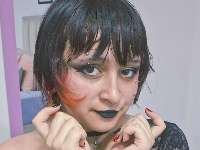 Nahaufnahme von MichellJula mit schwarzen Haaren, Pony und orangefarbenen Strähnen. Dunkles Make-up, schwarzer Lippenstift, Nasenring und Septum-Piercing. Choker mit Anhänger. Hände mit roten Nägeln, die das Gesicht umrahmen. Verdammt sexy..