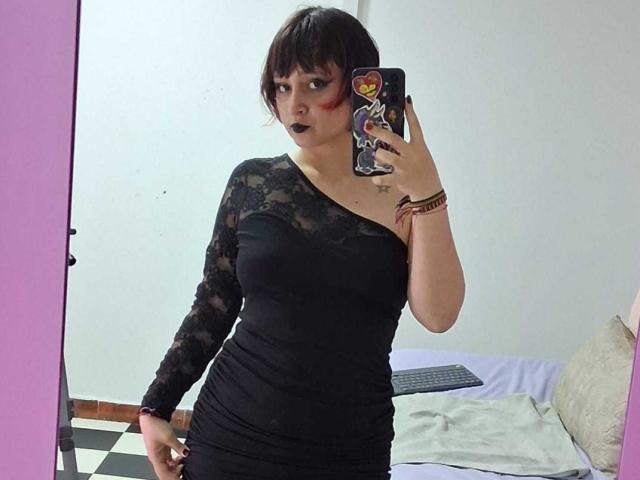 Photograph of MichellJula taking a mirror selfie. She's wearing a black, one-shoulder lace dress, looking at the camera with a slightly open mouth, holding her phone in her right hand. Background includes a bed with white and beige pillows. Her black hair is short and messy. She has a bracelet on her left wrist. Her phone case has a colorful sticker. The room has a pink door frame on the left. Her breasts are medium-sized and clearly visible under the dress. The lighting is bright, highlighting her facial features and outfit.