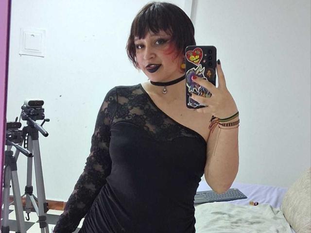 MichellJula, een vrouw met een goth-achtige uitstraling, met kort zwart haar en donkere lipstick, draagt een zwarte kanten jurk en een choker. Selfie in de spiegel. Ze houdt een telefoon vast met een kleurrijke hoes..