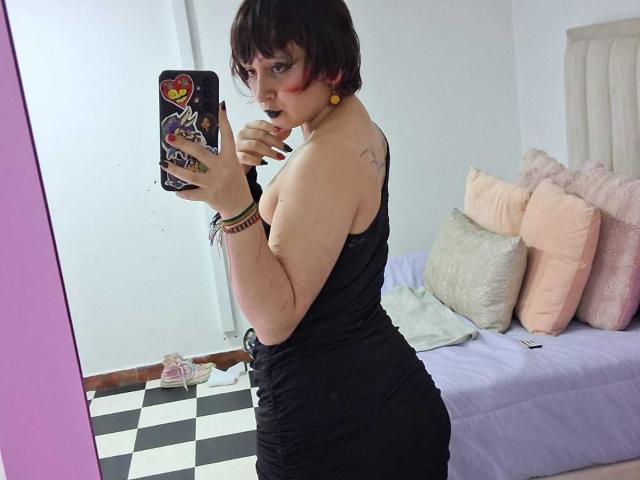 MichellJula, una donna formosa con capelli neri corti, rossetto nero e un vestito nero aderente, si scatta un selfie allo specchio. Lei tiene in mano un telefono con una custodia colorata. Pavimento a scacchi, cuscini rosa e beige. Assolutamente attraente..