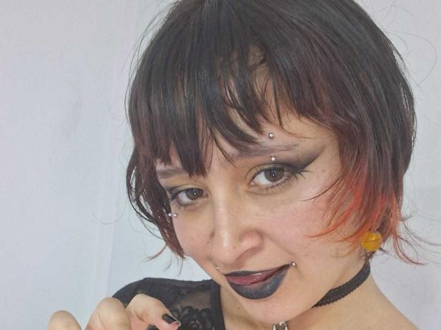 Photo en gros plan de MichellJula, avec des cheveux noirs courts, des pointes rouges, de multiples piercings au visage, du rouge à lèvres noir, et maquillage des yeux aux tons sombres.
elle porte un collier ras du cou noir et un haut en dentelle noire. Incroyablement sexy..