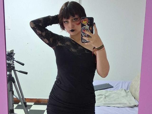 Autoportrait de MichellJula, debout, une main derrière la tête, portant un haut en dentelle noire, une jupe noire et un rouge à lèvres foncé. bracelets, tenant un téléphone avec une coque colorée. Lit blanc en arrière-plan. Incroyablement sexy..