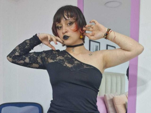 MichellJula, kurze, schwarze Haare mit roten Spitzen, schwarzes Langarm-Top aus Spitze, asymmetrisch geschnitten, schwarzer Lippenstift, schwarzer Choker. Stern-Tattoo auf der Brust, schwarze Nägel, Pose mit Händen in der Nähe des Gesichts, intensiver Blick, sexy..