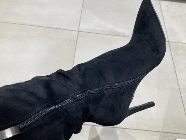 Close-up photo of DeesseBleue's foot wearing a black, high-heeled, suede boot with a silver zipper, angled upward, showing the boot's sharp point and metallic heel, against a white tiled floor.