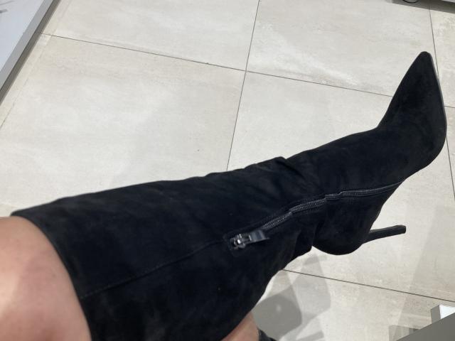 Primo piano della gamba sinistra di DeesseBleue, che indossa uno stivale nero in pelle scamosciata con un tacco alto e una cerniera. La scarpa ha la punta affusolata ed è posata su un pavimento lucido, rivestito di piastrelle bianche. Lo stivale è inclinato di un angolo..