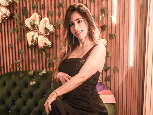 Victoria Jhonnes, com longos cabelos castanhos, vestindo um vestido preto de renda, sentada em um sofá verde estofado, sorrindo de forma sedutora, fundo de painel de madeira clara, flores artificiais, iluminação suave, tensão sexual..