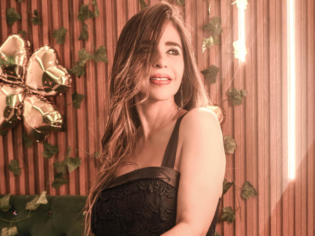 Fotografía de VictoriaJhonnes, cabello largo y castaño, vestido de encaje negro, sonriendo, ojos marrón claro, pared de madera a rayas, hiedra. y flores iluminadas en el fondo, luz suave, sensual, pose segura..