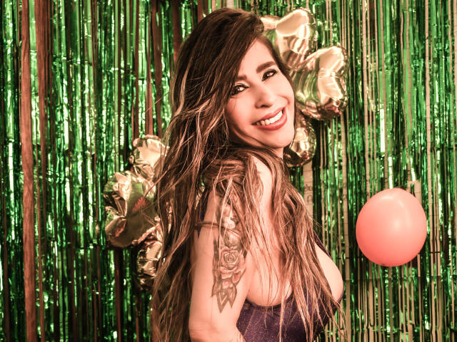 VictoriaJhonnes, mit langen, dunklen Haaren, die verführerisch lächelt, trägt ein glitzerndes Oberteil, vor einem grünen, metallischen Vorhang. Goldene Sternballons und ein roter Ballon im Vordergrund. Verdammt heiß.