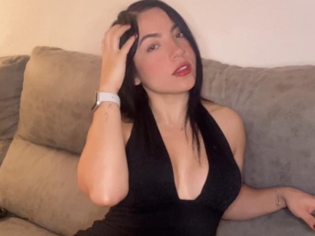 SexySussieX met zwart haar, rode lippenstift en een diepe zwarte haltertop, zittend op een beige bank, hand in haar, kijkend. verleidelijk, subtiele decolleté, horloge, ontspannen, casual uitstraling.