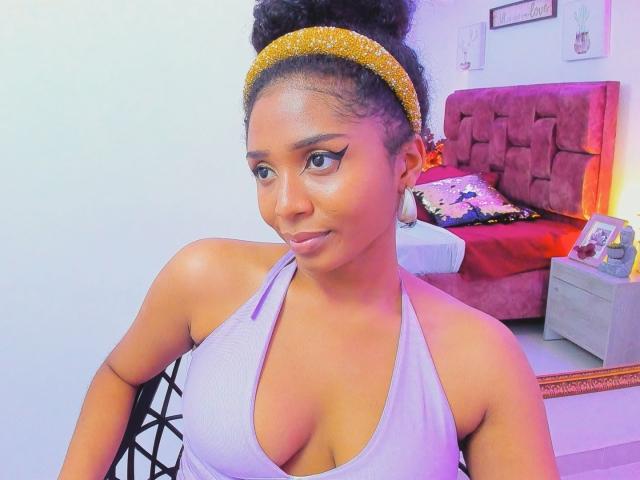 Photograph of MeganWlliams, dark-skinned woman with a high bun, wearing a white halter top and gold headband, showing cleavage, in a bedroom with a red bed and white walls. Fucking sexy.